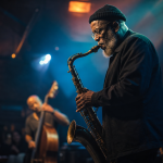 Joshua Redman tocando saxofone em apresentação de jazz com iluminação cênica azul e âmbar
