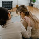 Duas mulheres sentadas em ambiente interno, conversando em um momento de apoio e empatia.