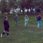 Crianças correm e brincam em um parque gramado cercado por árvores.