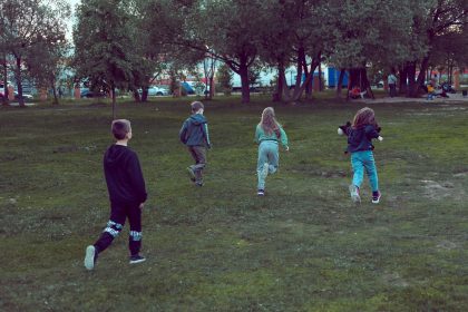 Crianças correm e brincam em um parque gramado cercado por árvores.