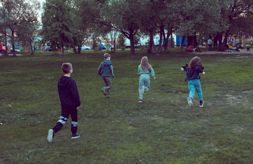 Crianças correm e brincam em um parque gramado cercado por árvores.