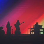 Músicos se apresentam em um palco iluminado por luzes coloridas diante do público.