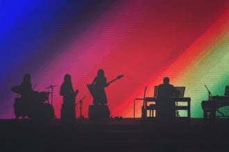 Músicos se apresentam em um palco iluminado por luzes coloridas diante do público.