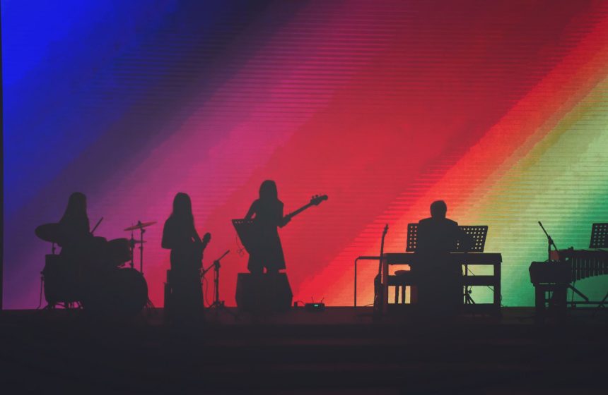 Músicos se apresentam em um palco iluminado por luzes coloridas diante do público.
